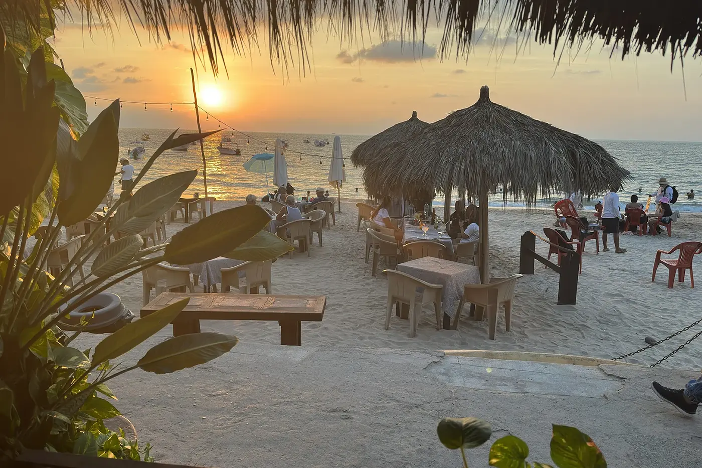 A beach at sunset with palm-leaf umbrellas and people dining.